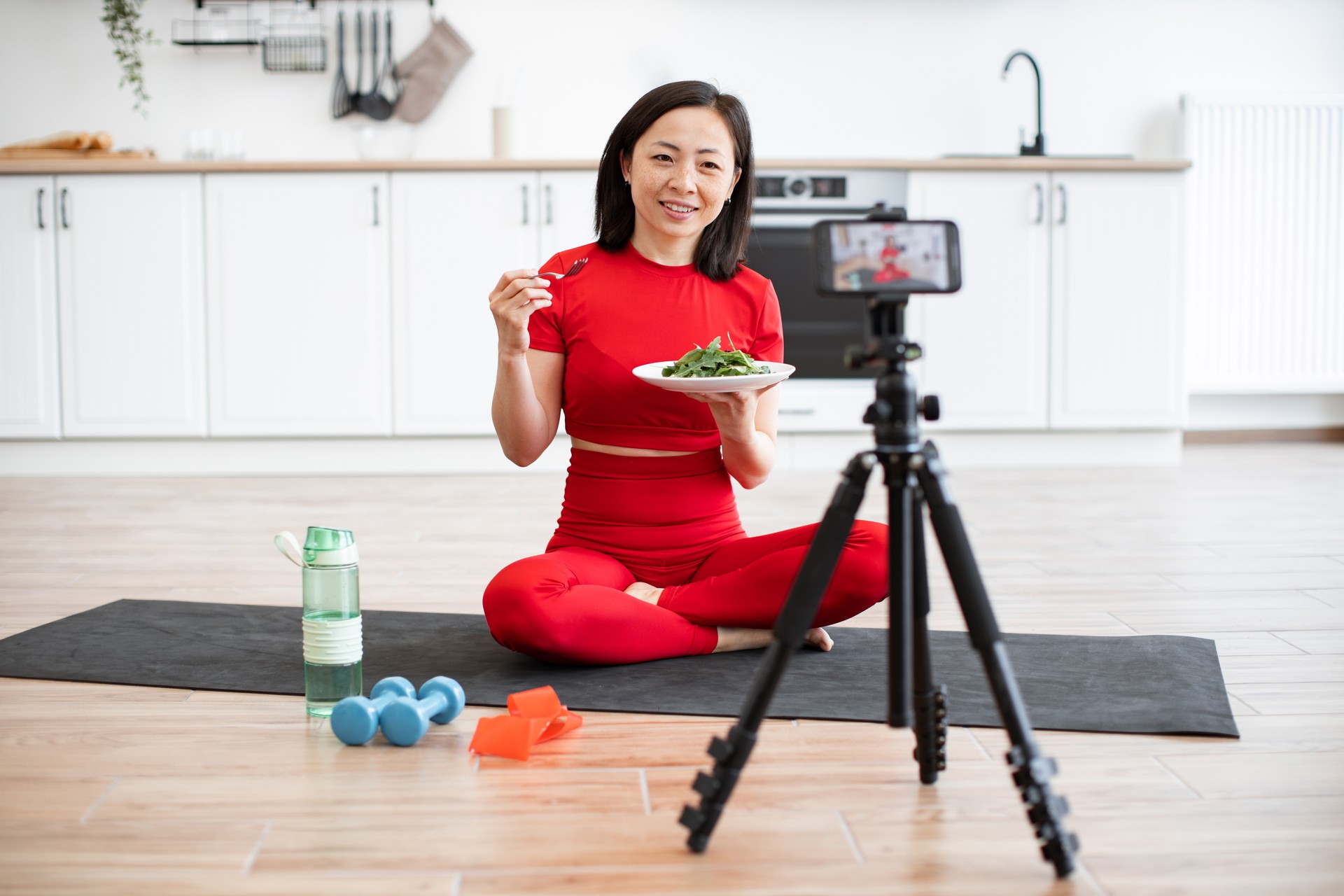 Mujer en ropa deportiva comiendo ensalada mientras graba un video de fitness en la cocina