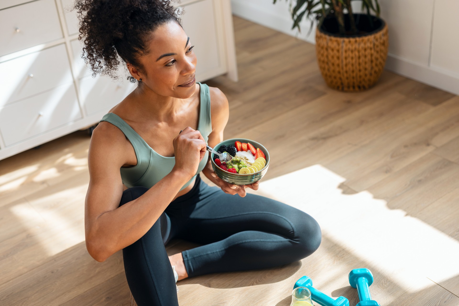 Mujer atlética comiendo un frutero saludable mientras está sentada en el suelo en la cocina de casa