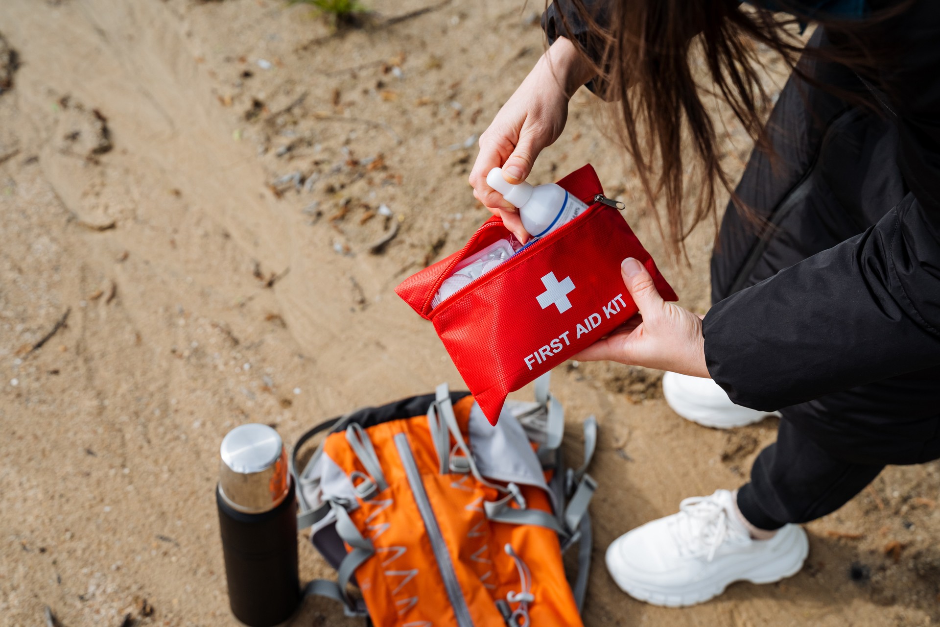 Garantice la preparación para la aventura al aire libre con un botiquín de primeros auxilios rojo, una mochila naranja, una botella de agua en terreno arenoso. Manténgase seguro haciendo senderismo, campamento, trekking con suministros de emergencia